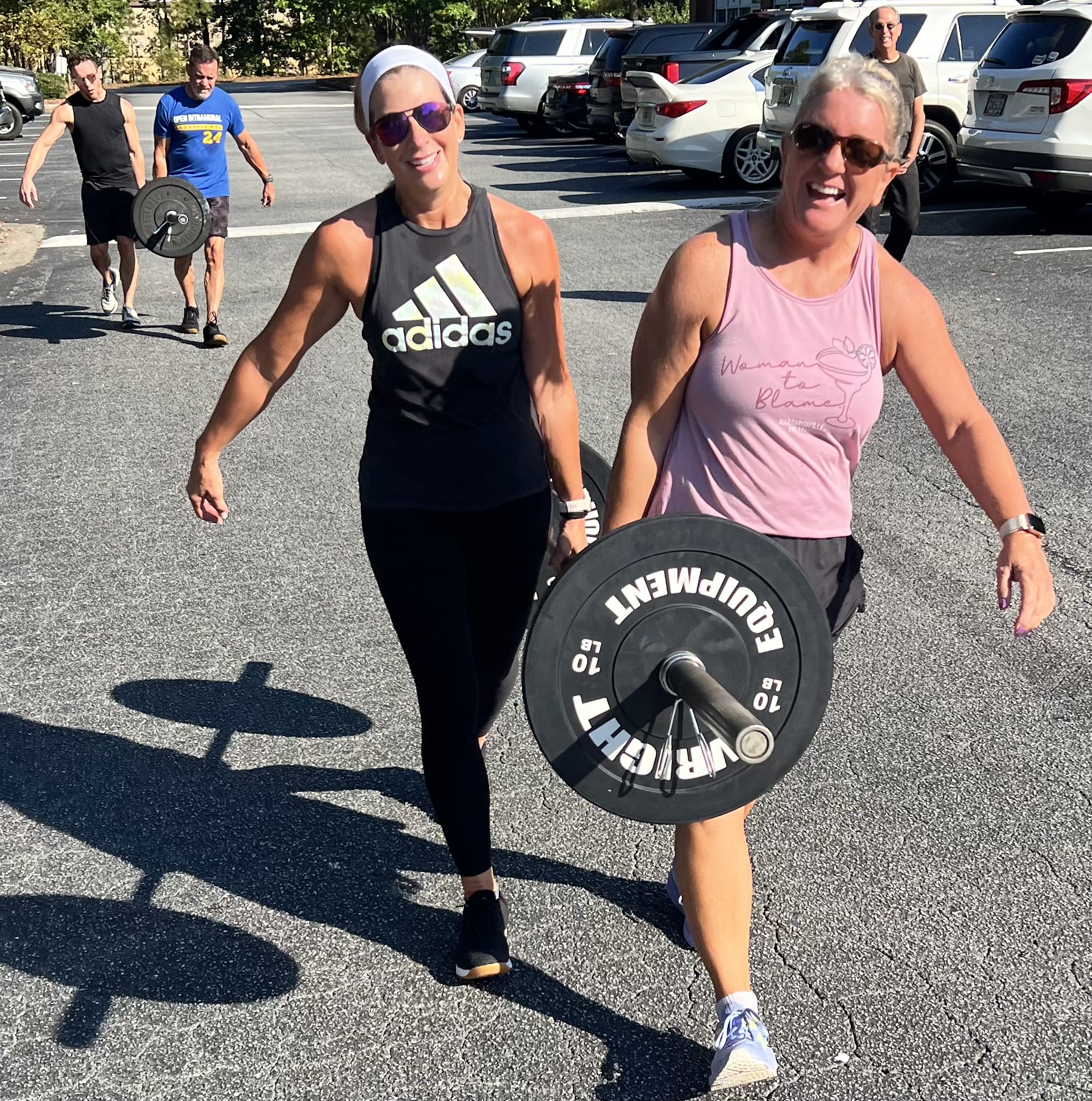 Two women smiling while carrying barbells during a CrossFit workout in the parking lot at CrossFit PTC.